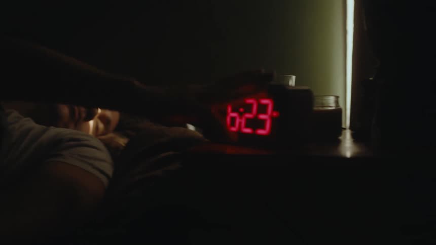 A man lying in bed presses a button on an alarm clock before going back to sleep