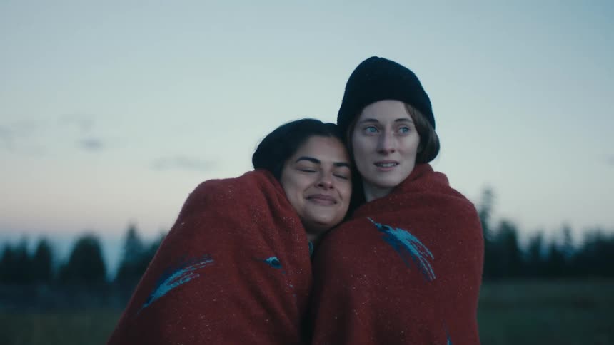 A pair of women standing in field while sharing a blanket embrace one another while looking into the distance