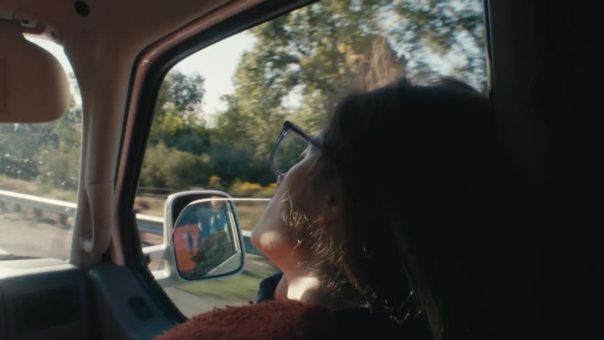 A woman rests with her eyes closed in the passenger seat of a vehicle as it travels through the countryside during the day