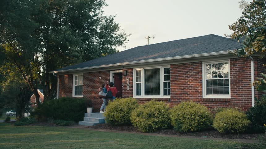 A pair of young woman approach the door of a house and are greeted with hugs from a woman residing within