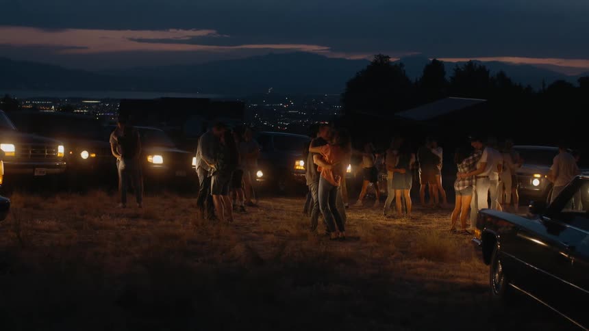 People dance in circle made of parked cars with lit headlights at night with city in the background