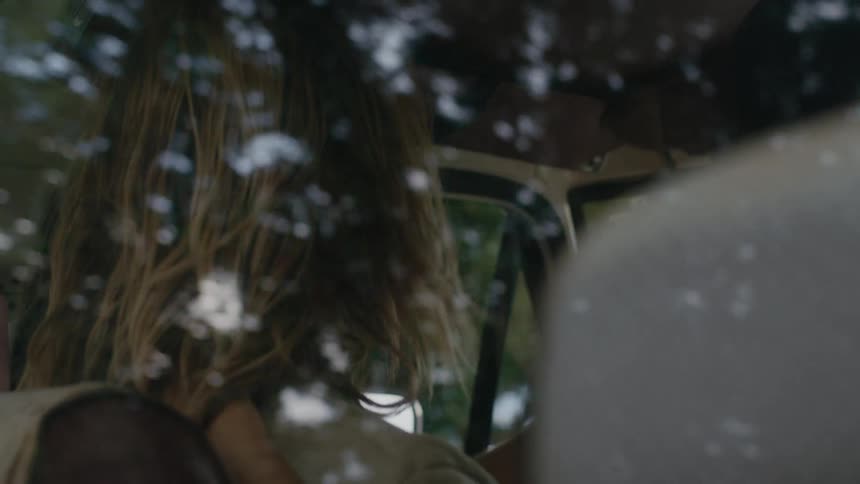A man with long blond hair and facial hair glances at his passenger while driving and chatting in his car