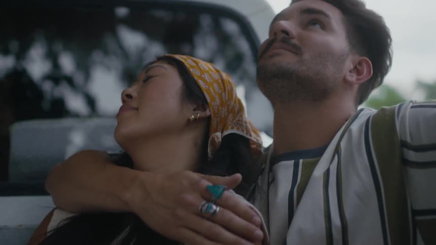 A man sits in the back of a driving truck with his arm around a woman and kisses her on the face and she holds his arm and leans her head on him
