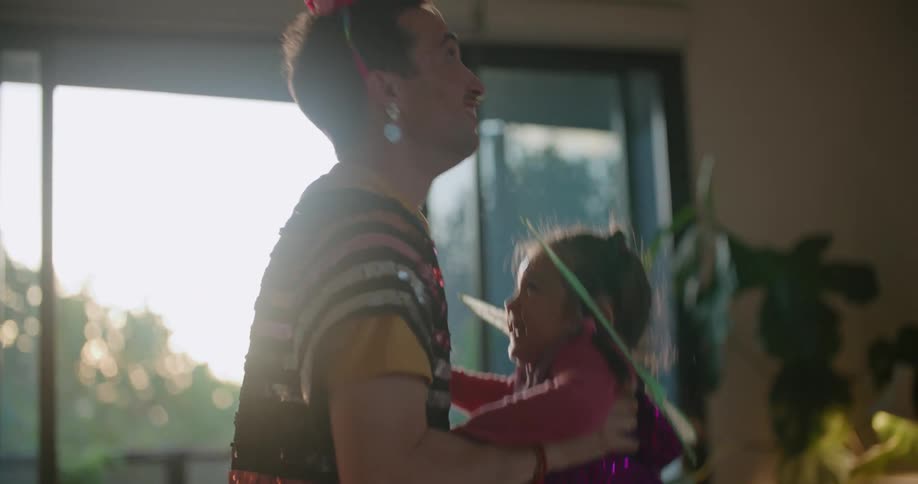 A man is wearing a sequined shirt and a party hat and lifting his young daughter wearing fairy wings into the air