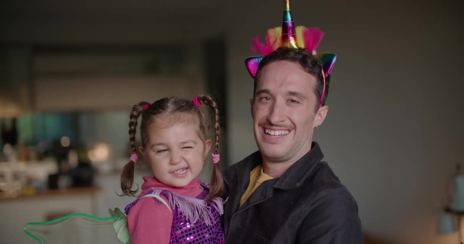 A man is wearing a party hat and holding his young daughter who is wearing green fairy wings and a purple sequined shirt