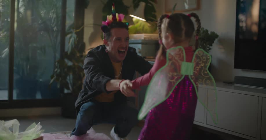 A man is playing with his young daughter who is wearing green fairy wings and he rolls on the floor and holds her in the air