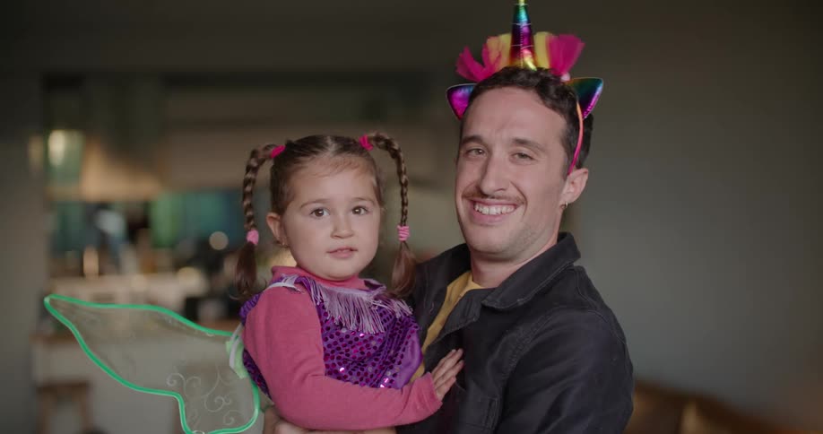 A man is wearing a party hat and holding his young daughter who is wearing green fairy wings and a purple sequined shirt