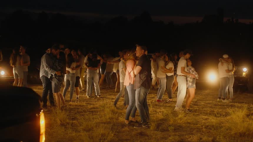 A group of people slow dance in a field illuminated by vehicle headlights
