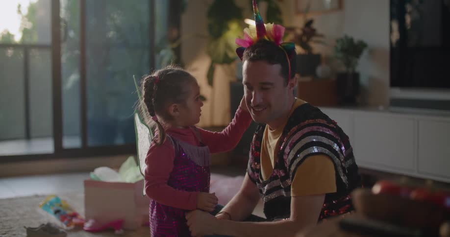 A man is wearing a sequined shirt and a party hat and sitting on the floor playing with his young daughter wearing fairy wings