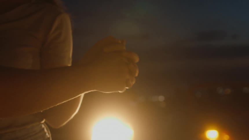 A couple are outside dancing and their hands are clasped together as a light shines brightly behind them