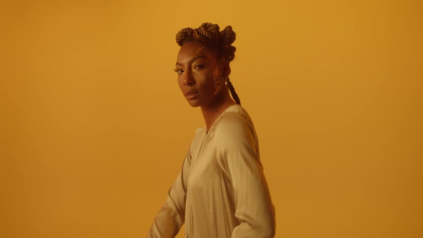 A woman kneeling before an orange background slowly turns her head to look into the camera