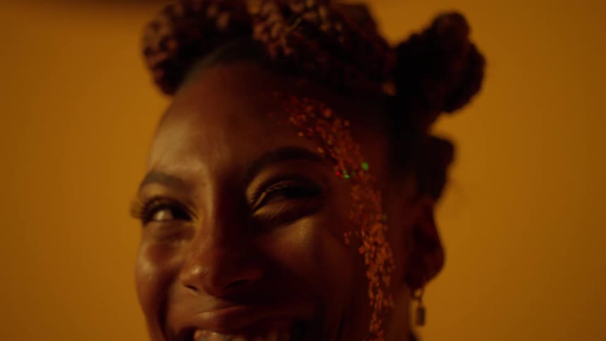 A black woman with glitter on her face is standing near a bright yellow wall smiling and laughing as she looks around