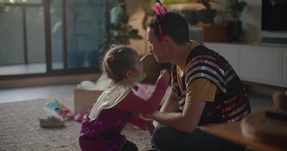 A man is wearing a sequined shirt and a party hat and sitting on the floor playing with his young daughter wearing fairy wings