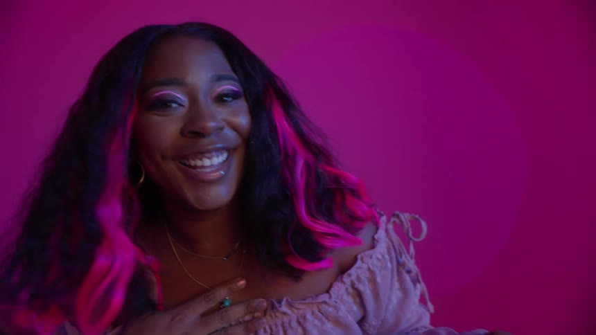 A woman with pink streaks in her hair poses in a studio with pink walls