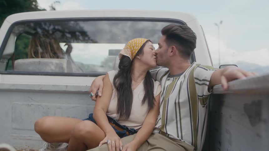 A man and a woman are sitting in the back of a pickup truck as it travels on a road and they kiss