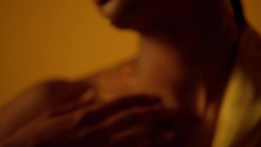 A young woman with glitter on her face and body runs her hand down her face and on to her chest as she caresses her body