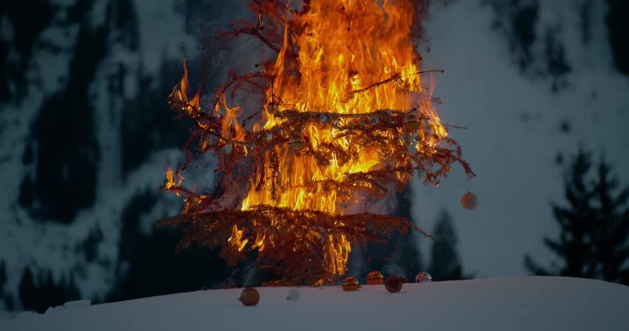 An outdoor christmas tree is on fire and ornaments are on the snow covered ground