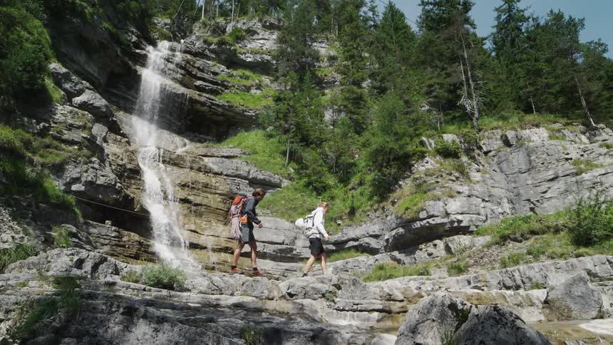 A man and woman walk on stones to cross a stream below a waterfall on their hike