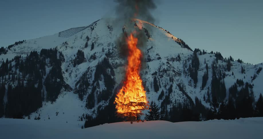 A christmas tree is in flames in front of a snow covered mountain and ornaments are falling from the branches
