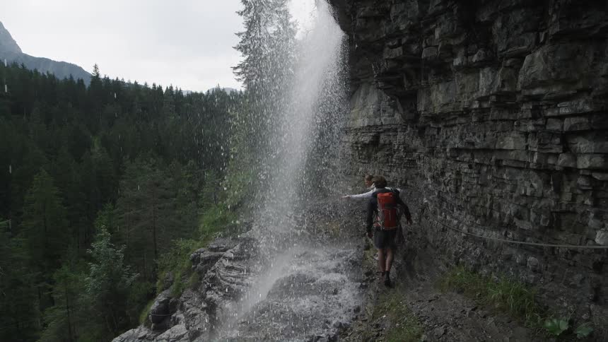 Two people are walking along a ledge as waterfalls splash down in front of them