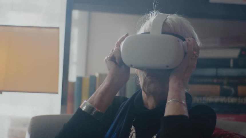 An elderly woman sitting indoors puts a virtual reality headset on her head and exclaims at what she sees