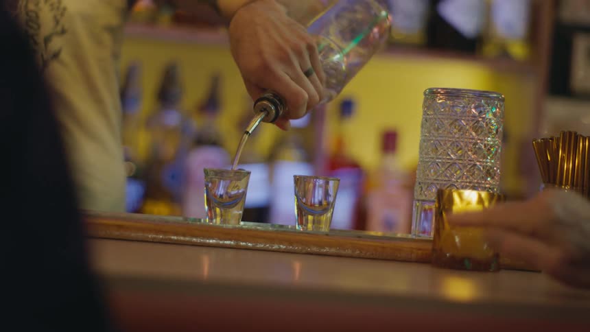 A bartender pours two shots and serves them to customers sitting at the bar