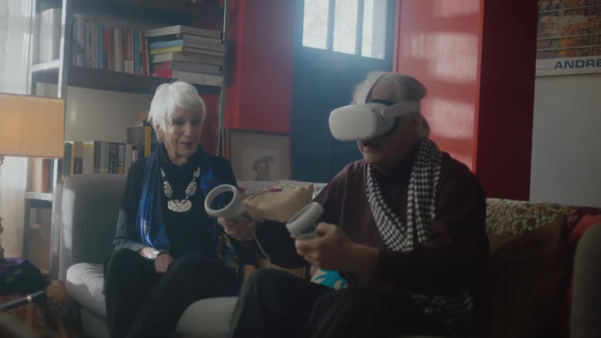 A man and a woman are sitting on a couch and he puts on a virtual reality headset and picks up the controllers and moves them around