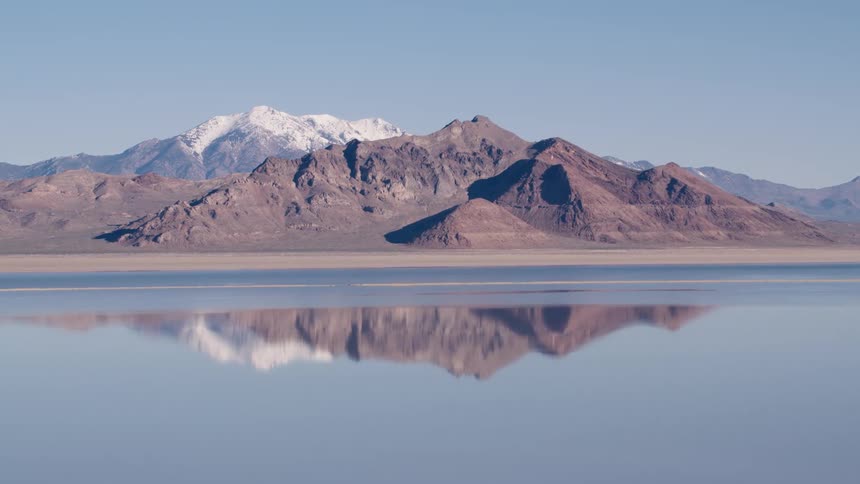 A mountain range reflects in the water and snow covered mountains are in the distance under a clear sky