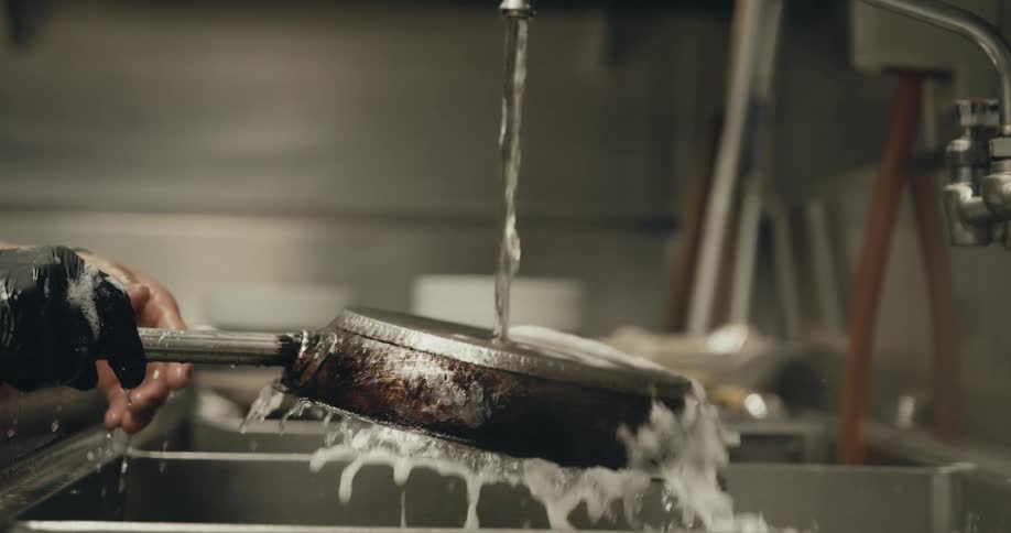 A dishwasher cleaning an iron pan under a stream of water on a kitchen sink in a restaurant