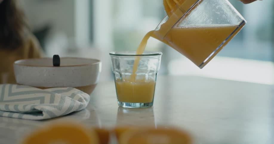 A person pours orange juice from a pitcher into a drinking glass on a kitchen table