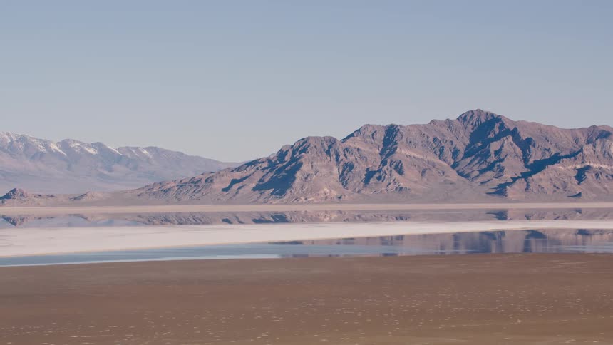 A mountain range reflects in the water and more mountains are in the distance under a clear sky
