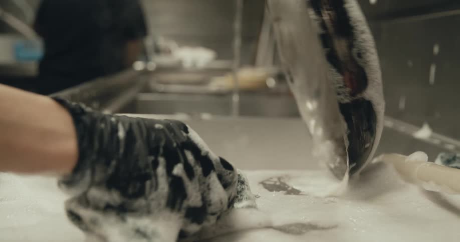 A man washes a pan at the restaurant sink as a busboy empties a tray behind him