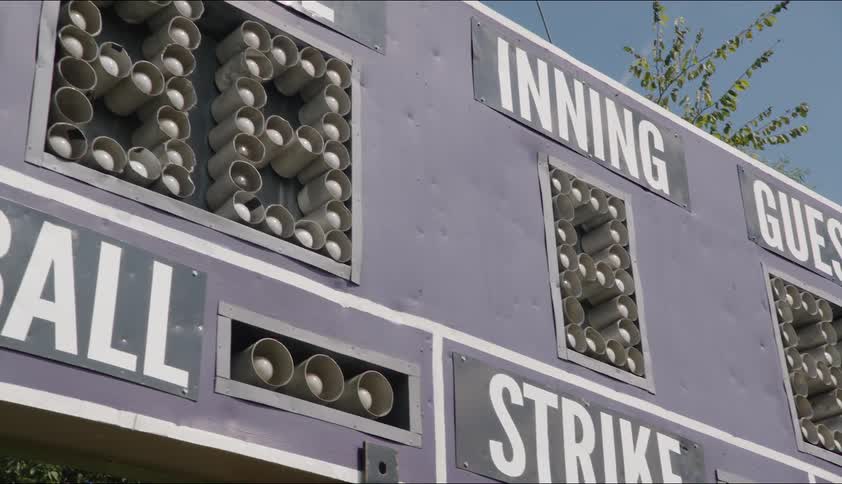 A baseball scoreboard with trees in the distance under a clear blue sky