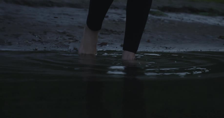 A barefoot person walks over a muddle shore into a lake