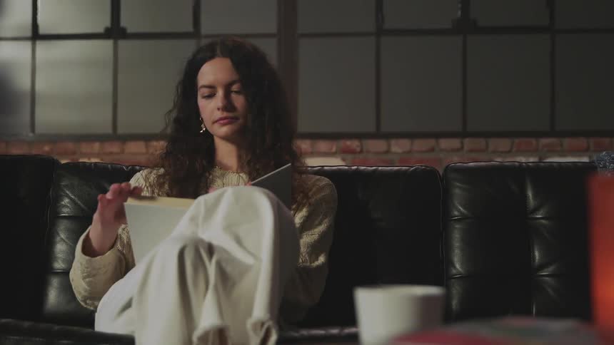 Woman wearing sweater sits on couch with hardback book and flips through pages