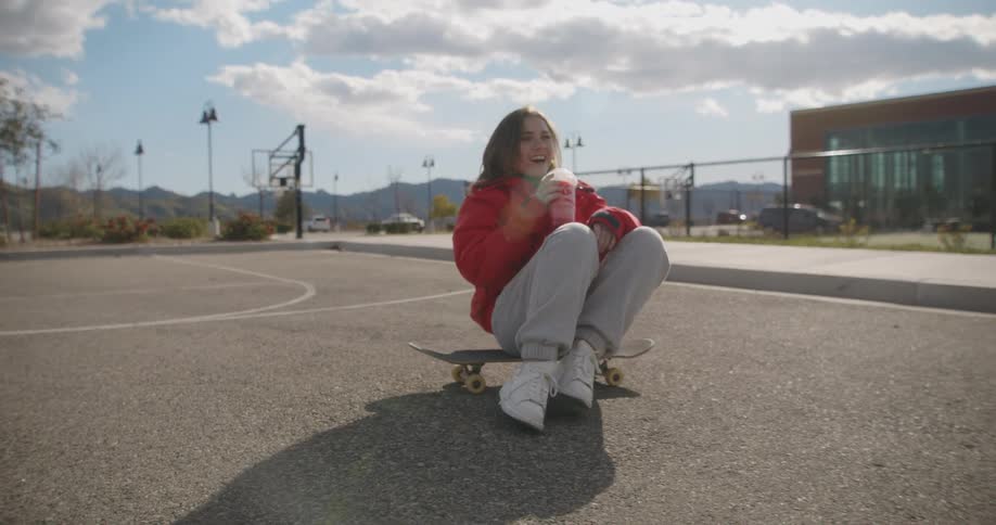 A woman drinks from a plastic cup while sitting on a skateboard on an outdoors basketball court during the day