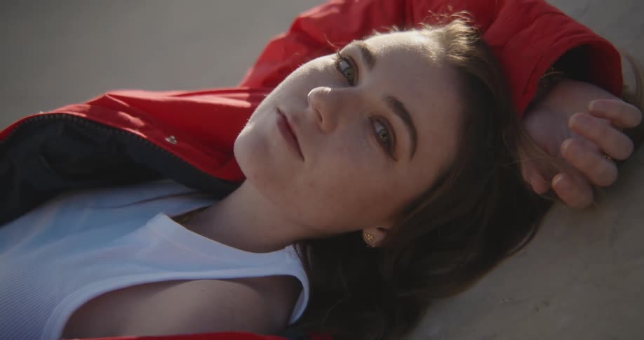 A teenage girl wearing a red jacket is laying on the concrete and she turns her head and smiles