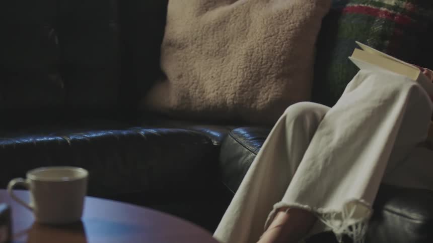 A woman places a coffee cup on a table before sitting on a sofa and beginning to read a book