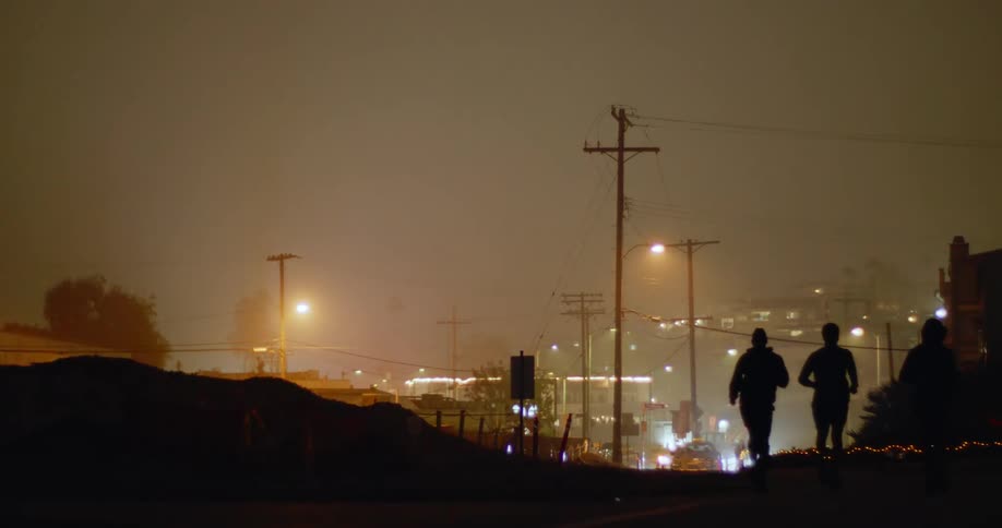 Four men run together outside as car headlights glare in the distance