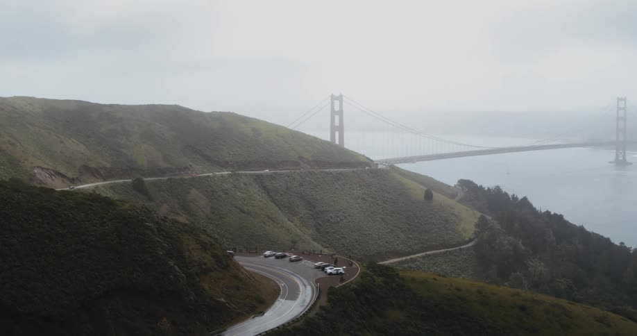 Cars are in a parking lot and other automobiles travel on a road leading to a suspension bridge stretching across the water and into the fog on the other side