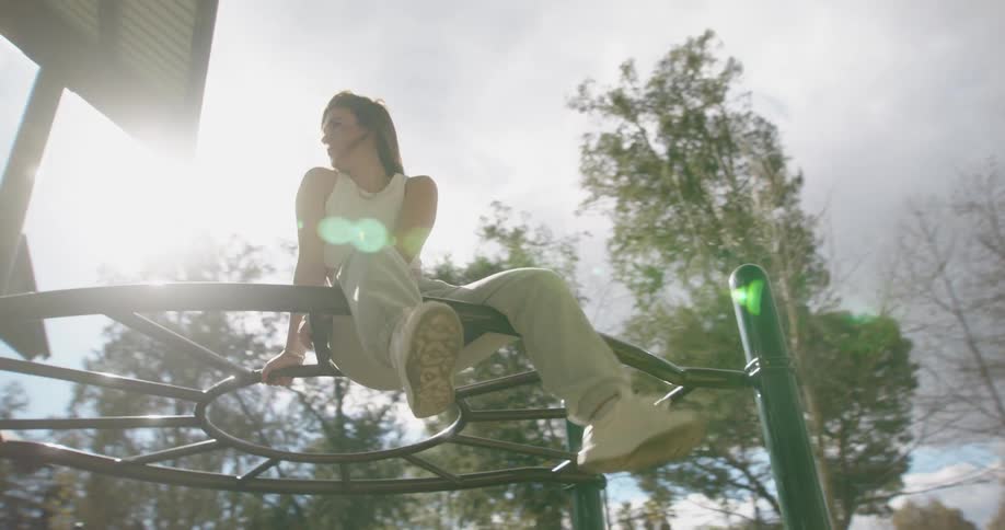 A young woman kicks her legs while sitting atop the monkey bars on a playground during the day