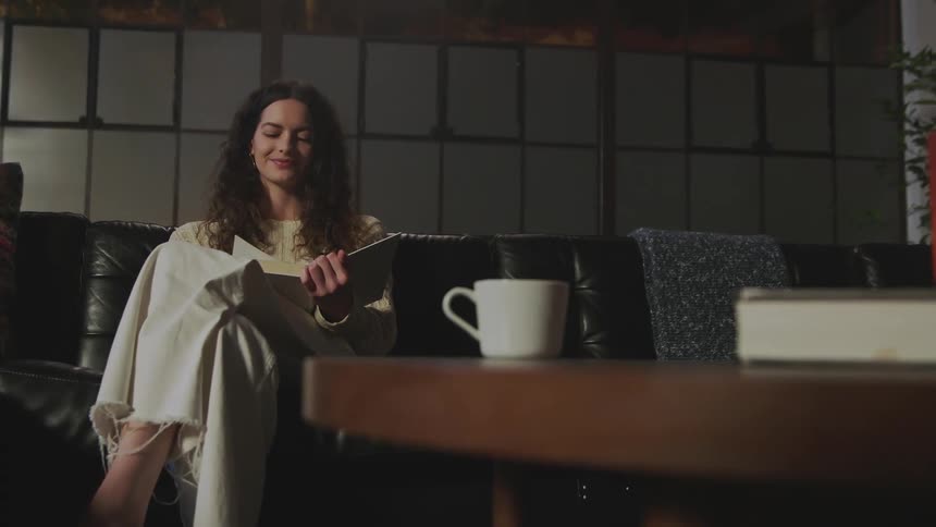 A woman is sitting on a couch reading a book and there is a coffee cup on the table near her