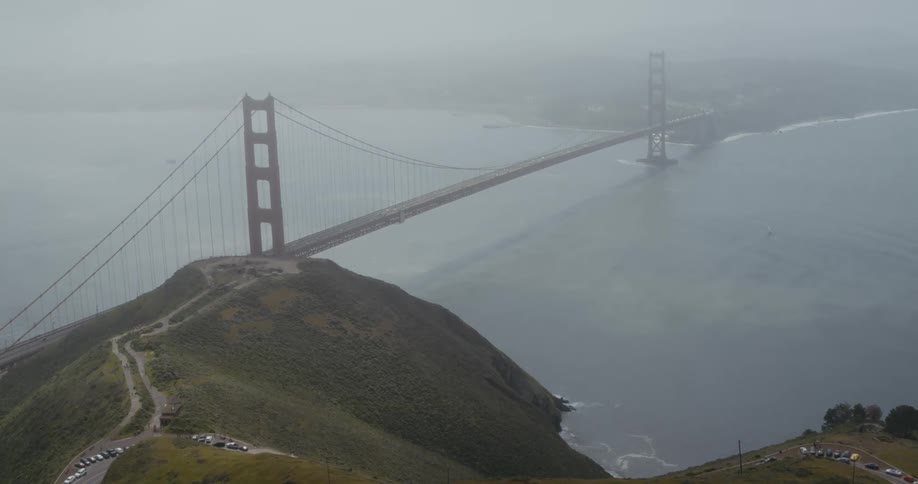 A road along a hilltop leads to a suspension bridge stretching across the water and into the fog
