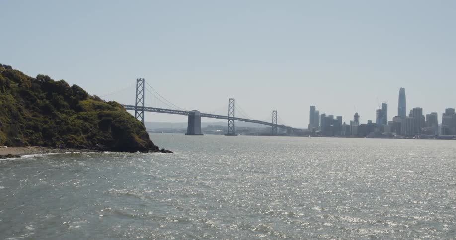Water sparkles as it moves and there is a suspension bridge leading to a big city with tall buildings and skyscrapers