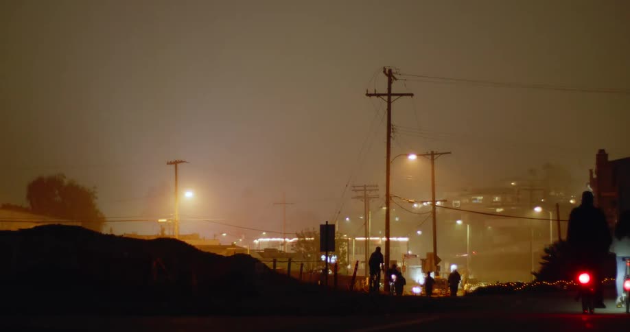 Pedestrians cyclists and vehicles move along road silhouetted against streetlights on foggy night