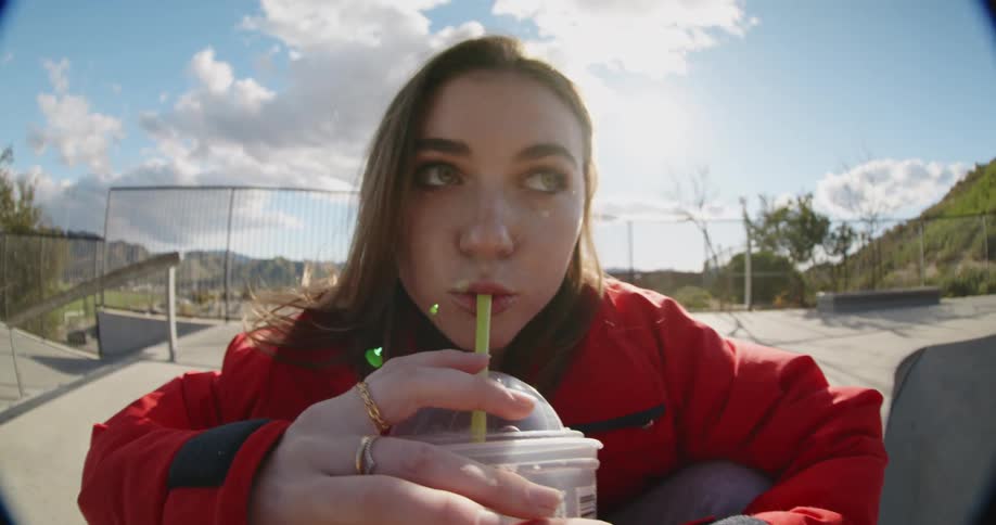 A woman sipping a drink through a straw while at a skate park on a bright sunny day