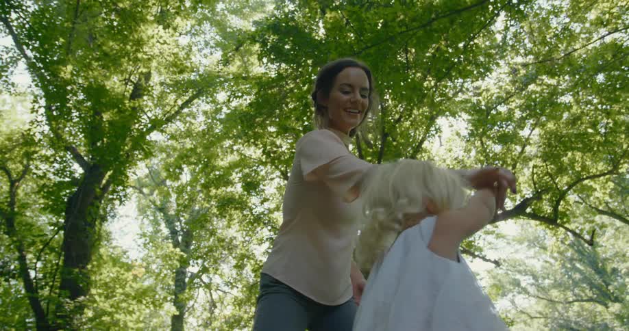 Mother twirls girl then crouches next to her as they stand under the trees