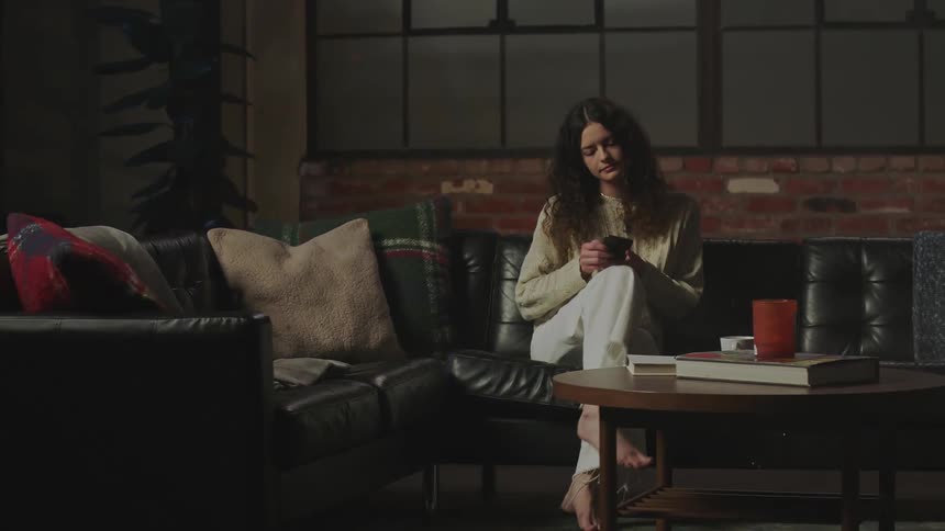 A woman at home walks to the couch and sits down and looks at her cell phone and scrolls on it