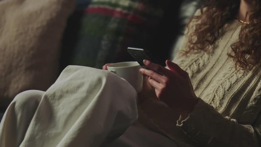 A woman is sitting on a couch holding a coffee cup and using her cell phone