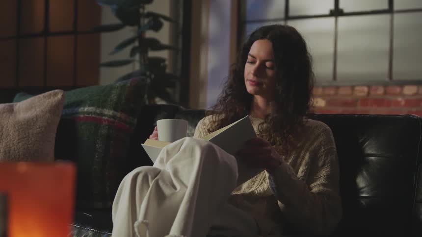 A woman holding a coffee cup reads a book while sitting on a leather sofa
